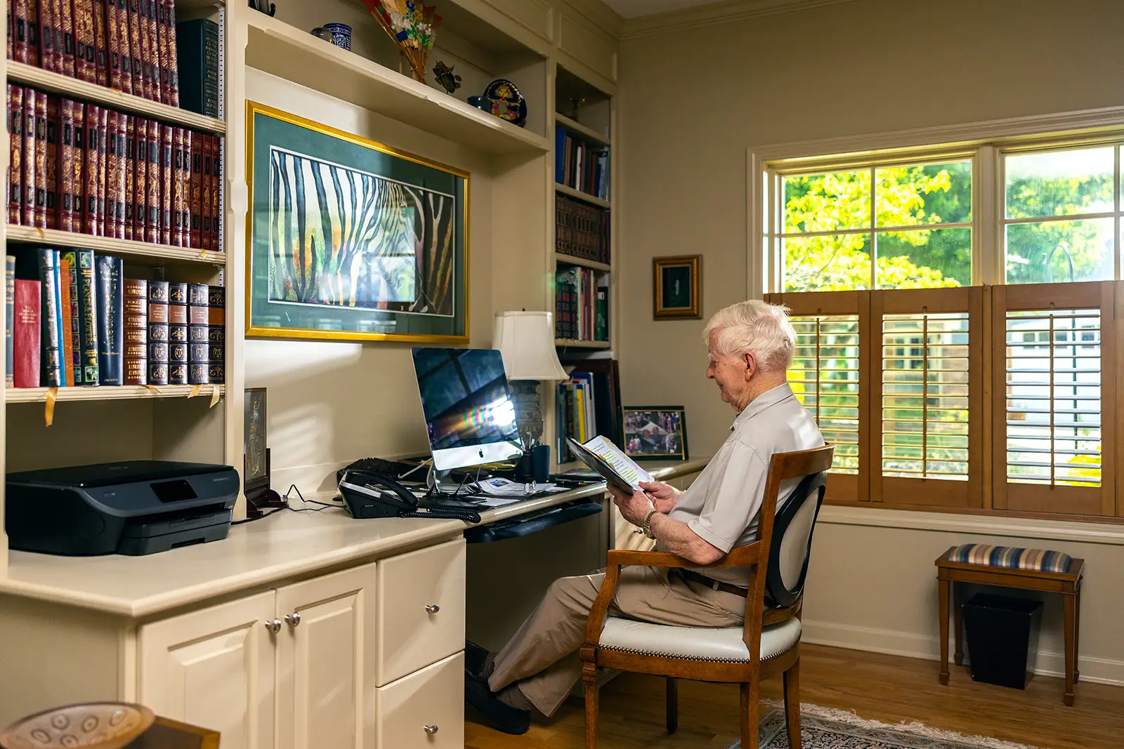 A senior resident at Essex Meadows sits in a home study surrounded by bookshelves, reading a booklet in front of a computer desk while engaging in daily cognitive activities and brain games for seniors.