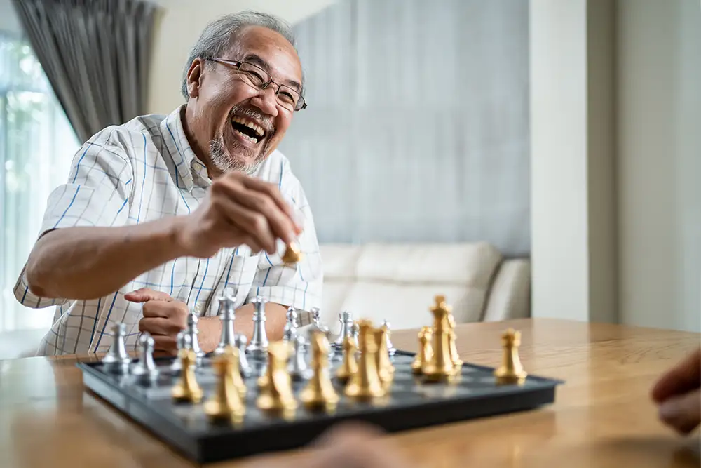 A senior at Essex Meadows laughs while making a move on a metallic chessboard, enjoying brain games for seniors with a friend.