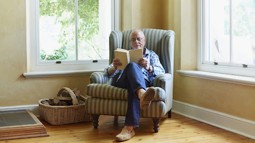 Full length of senior man reading books on aging well while relaxing on chair at home